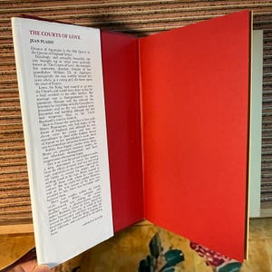 Puede incluir: Libro abierto titulado "The Courts of Love" de Jean Plaidy. La portada del libro es blanca con texto negro, y las p&aacute;ginas interiores son blancas con texto negro. La contraportada es roja. El libro est&aacute; abierto en una p&aacute;gina de texto.