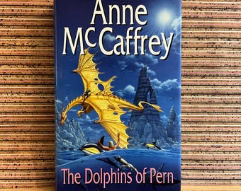 May include: A hardcover book titled "The Dolphins of Pern" by Anne McCaffrey. The cover features a golden dragon flying over water with dolphins and a person. The background is a blue sky with a bright sun and rocky formations.
