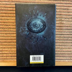 May include: A dark blue book cover with a silver eye design and a white ISBN barcode. The text on the barcode reads "ISBN 0-385-60615-X".