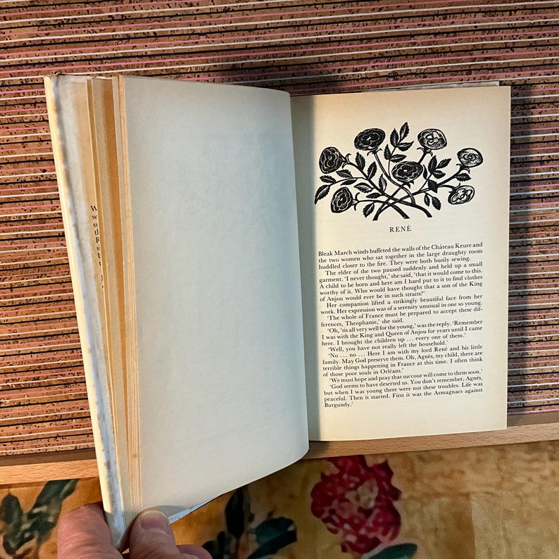 Puede incluir: Un libro abierto con texto y una ilustraci&oacute;n en blanco y negro de rosas. El libro est&aacute; sobre una superficie de madera. El texto incluye la palabra "RENE" y un pasaje sobre un ni&ntilde;o y el rey de Anjou.