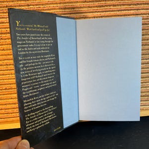 May include: A close-up of the back cover of a fantasy novel titled "The Golem's Eye". The cover features a dark blue background with gold lettering and a detailed illustration of a city skyline. The text on the cover reads "Two years have passed since the events of The Amulet of Samarkand, and the young magician Nathaniel is fast rising through the government ranks. It is up to him to put an end to the thefts and raids inflicted on London by the mysterious Resistance."