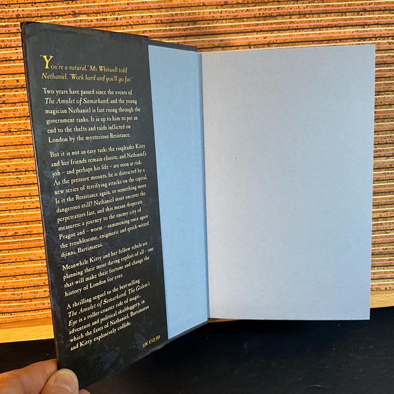 May include: A close-up of the back cover of a fantasy novel titled "The Golem's Eye". The cover features a dark blue background with gold lettering and a detailed illustration of a city skyline. The text on the cover reads "Two years have passed since the events of The Amulet of Samarkand, and the young magician Nathaniel is fast rising through the government ranks. It is up to him to put an end to the thefts and raids inflicted on London by the mysterious Resistance."