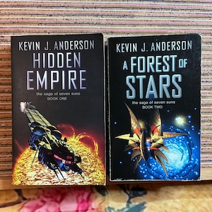 Könnte beinhalten: Zwei Science-Fiction-Romane von Kevin J. Anderson. "Hidden Empire" zeigt ein Raumschiff über einer Sonnengrafik. "A Forest of Stars" zeigt ein Sternenfeld und ein Raumschiff. Beide Bücher gehören zu "the saga of seven suns".