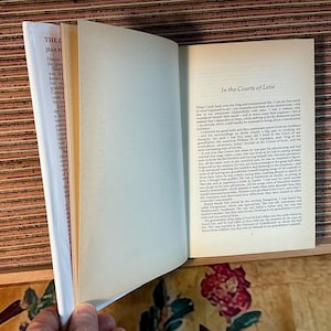 Puede incluir: Un libro abierto con el t&iacute;tulo "In the Courts of Love" visible en la p&aacute;gina derecha. La p&aacute;gina izquierda muestra texto de "The Queen's Dazzle" de Jean Plaidy. El libro est&aacute; sobre una superficie de madera.