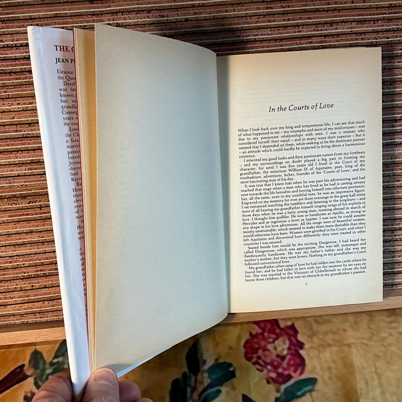 Puede incluir: Un libro abierto con el t&iacute;tulo "In the Courts of Love" visible en la p&aacute;gina derecha. La p&aacute;gina izquierda muestra texto de "The Queen's Dazzle" de Jean Plaidy. El libro est&aacute; sobre una superficie de madera.
