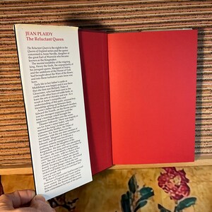 Puede incluir: Libro abierto titulado "The Reluctant Queen" de Jean Plaidy. La portada del libro es blanca con texto negro, y las p&aacute;ginas interiores son rojas. El libro est&aacute; abierto, revelando texto sobre la reina de Inglaterra.