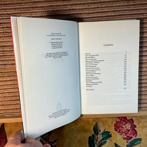 Puede incluir: Un libro abierto con lomo rojo, que muestra la p&aacute;gina de contenido e informaci&oacute;n de derechos de autor. Se ven t&iacute;tulos de cap&iacute;tulos como "The Long Betrothal" y "Married Bliss". El libro est&aacute; sobre una superficie de madera.