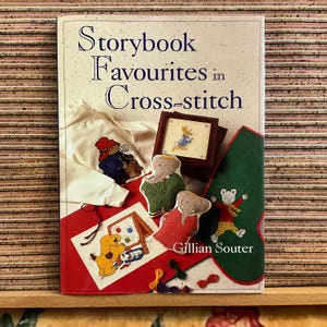 May include: A book titled "Storybook Favourites in Cross-stitch" featuring cross-stitch designs. The image shows a framed cross-stitch of a rabbit, stuffed elephant toys, and a cross-stitch of a dog painting. The cover displays a white sweatshirt with a cross-stitch design.