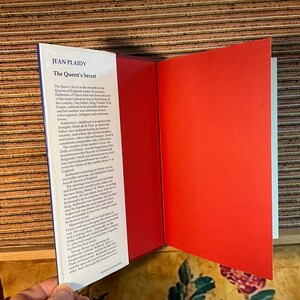 Puede incluir: Un libro abierto titulado "The Queen's Secret" de Jean Plaidy. Las p&aacute;ginas del libro son rojas y blancas, con texto en la p&aacute;gina izquierda. El libro forma parte de la serie Queens of England.