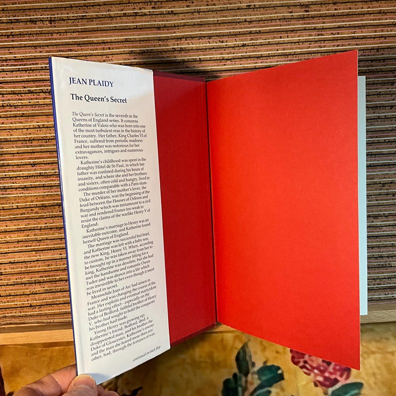 Puede incluir: Un libro abierto titulado "The Queen's Secret" de Jean Plaidy. Las p&aacute;ginas del libro son rojas y blancas, con texto en la p&aacute;gina izquierda. El libro forma parte de la serie Queens of England.