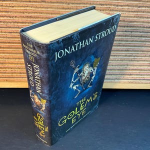 May include: A hardcover book titled "The Golem's Eye" by Jonathan Stroud. The book is part of the "Bartimaeus Trilogy". The cover features a blue background with a skeletal figure holding a sword and a mask.