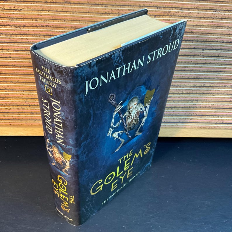 May include: A hardcover book titled "The Golem's Eye" by Jonathan Stroud. The book is part of the "Bartimaeus Trilogy". The cover features a blue background with a skeletal figure holding a sword and a mask.