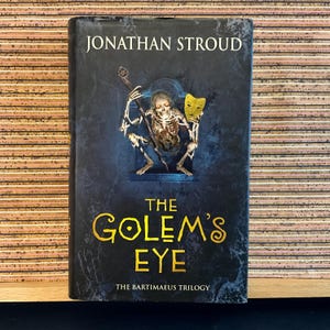 May include: A book cover with a dark blue background featuring a skeletal figure holding a staff and a yellow mask. The title "The Golem's Eye" is written in gold lettering with a black circle in the "O" of "Golem". The text "The Bartimaeus Trilogy" is written in white lettering at the bottom of the cover.