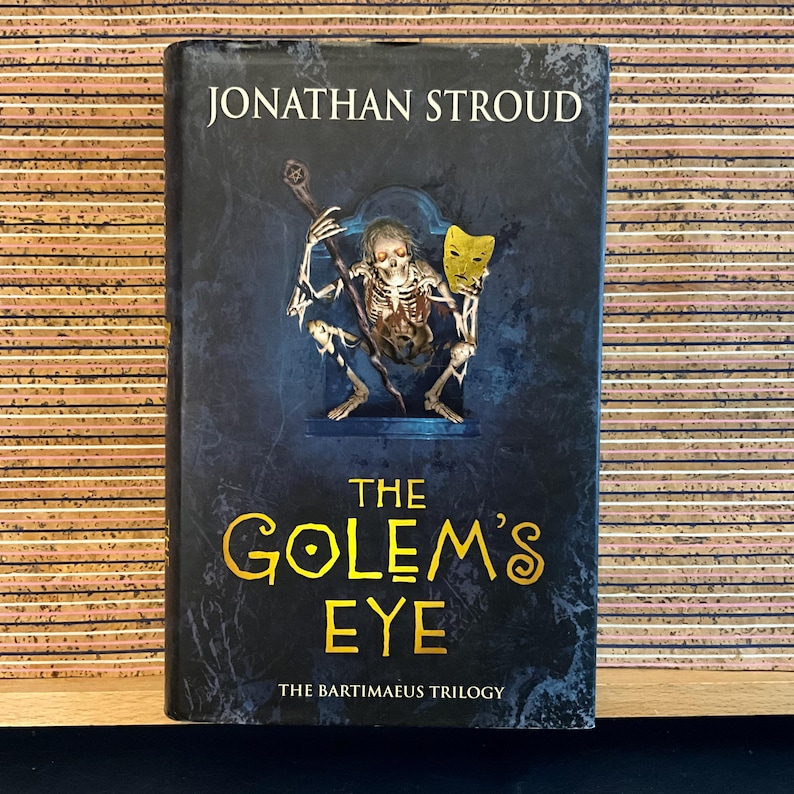 May include: A book cover with a dark blue background featuring a skeletal figure holding a staff and a yellow mask. The title "The Golem's Eye" is written in gold lettering with a black circle in the "O" of "Golem". The text "The Bartimaeus Trilogy" is written in white lettering at the bottom of the cover.