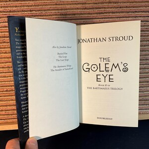 May include: The Golem's Eye, Book II of The Bartimaeus Trilogy by Jonathan Stroud. The book cover features a white background with black text and a stylized illustration of an eye.