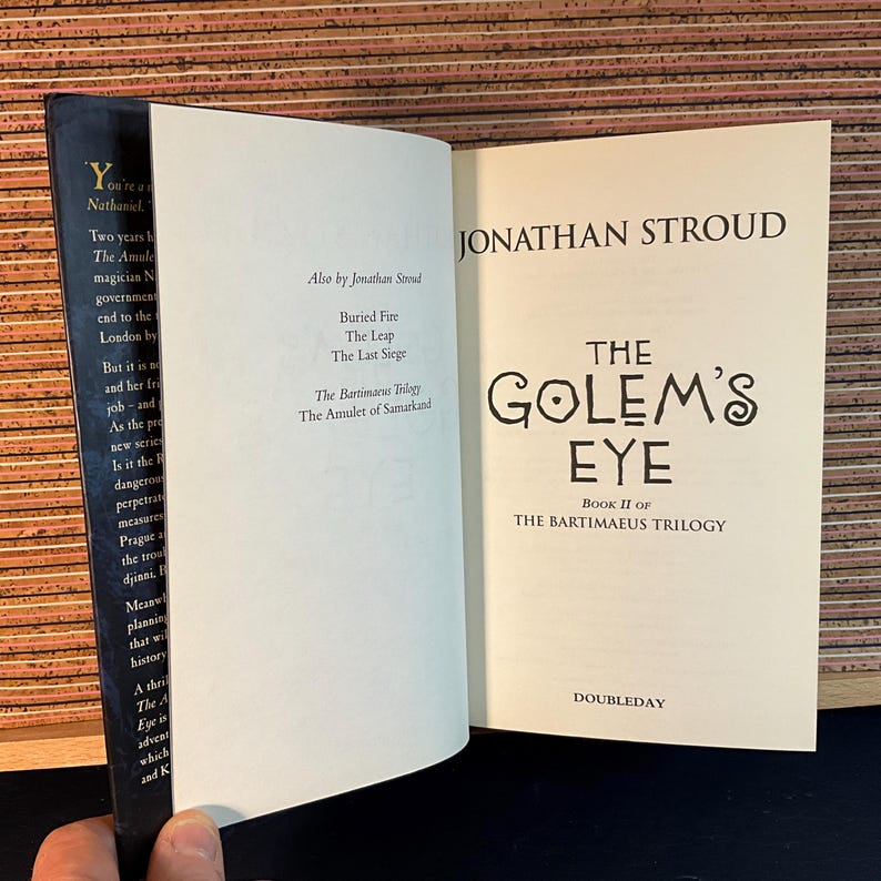 May include: The Golem's Eye, Book II of The Bartimaeus Trilogy by Jonathan Stroud. The book cover features a white background with black text and a stylized illustration of an eye.