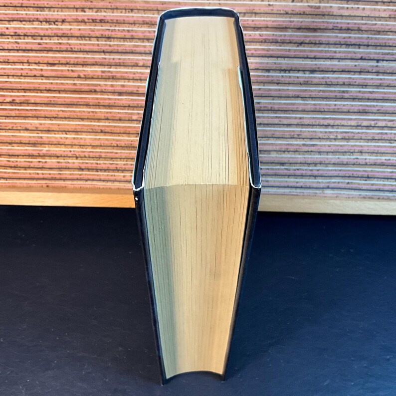 May include: A black hardcover book with a cream-colored spine. The book is standing upright on a dark surface.