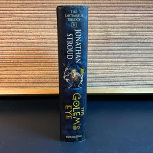 May include: The Golem's Eye by Jonathan Stroud, book two in The Bartimaeus Trilogy, with a dark blue cover and gold lettering. The cover features a skeletal figure holding a key.