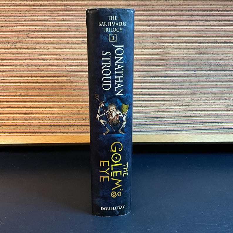 May include: The Golem's Eye by Jonathan Stroud, book two in The Bartimaeus Trilogy, with a dark blue cover and gold lettering. The cover features a skeletal figure holding a key.