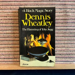 May include: A paperback book cover with a black background and the title "The Haunting of Toby Jugg" by Dennis Wheatley. The cover features a woman in a fiery setting with a silver bowl and a black spider.