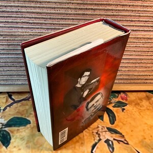 May include: A hardcover book with a red cover featuring a black and white image of a man wearing glasses. The book's pages are cream-colored, and a white bookmark is inserted. The book is on a floral patterned surface.