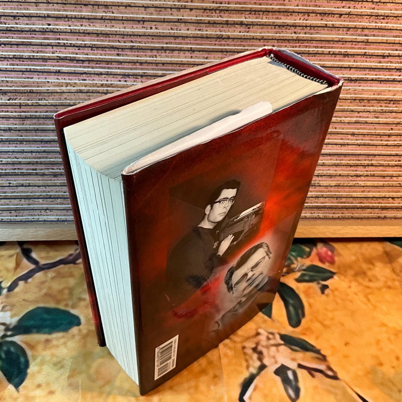 May include: A hardcover book with a red cover featuring a black and white image of a man wearing glasses. The book's pages are cream-colored, and a white bookmark is inserted. The book is on a floral patterned surface.