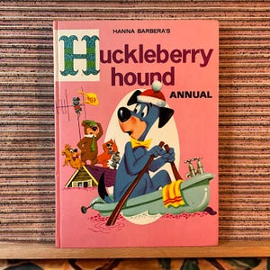 Könnte beinhalten: Ein Vintage-Huckleberry-Hound-Jahresbuch mit rosa Einband. Das Cover zeigt ein Cartoon-Bild von Huckleberry Hound in einem Boot, zusammen mit anderen Cartoon-Figuren. Der Titel ist in großen, kastanienbraunen Buchstaben.