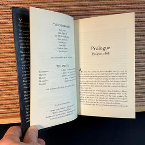 May include: An open book with a brown leather cover. The book is titled "The Commoners" and "The Spirits". The text on the page describes the characters and spirits in the book. The book is open to the prologue, which is set in Prague in 1868.