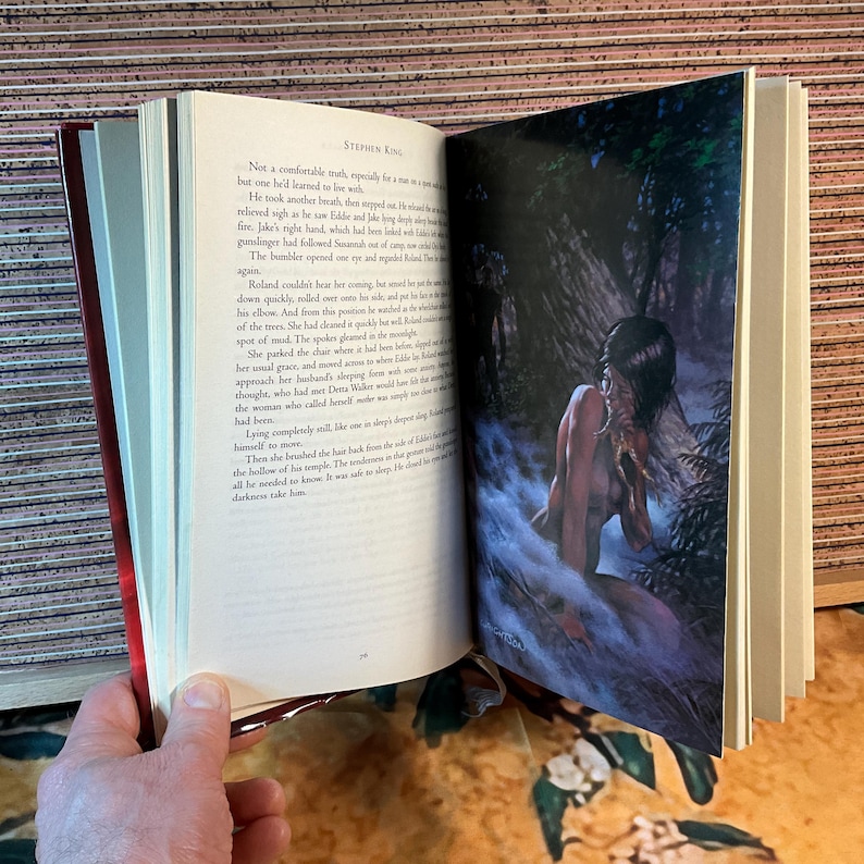 May include: An open book with text and an illustration. The illustration depicts a person in a misty, wooded area. The book's pages are cream-colored, and the cover is red. The text on the page is from Stephen King.