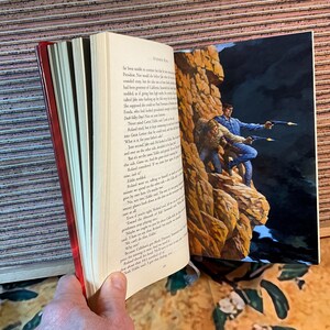 May include: Open book with a red spine, featuring text and a color illustration of two men with guns on a rocky cliff. The illustration is on the right page, while the left page displays printed text.