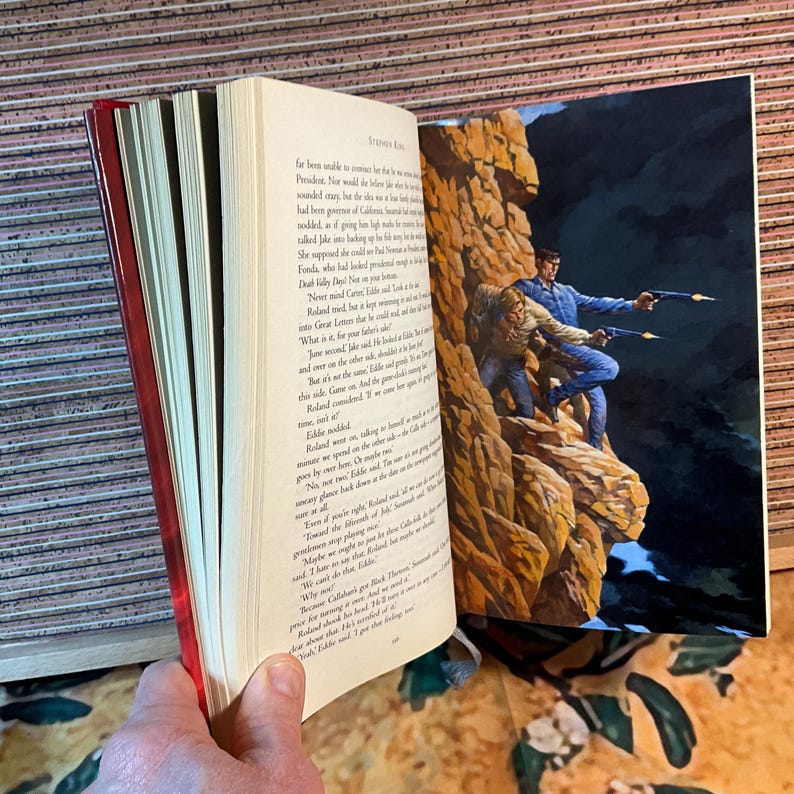 May include: Open book with a red spine, featuring text and a color illustration of two men with guns on a rocky cliff. The illustration is on the right page, while the left page displays printed text.