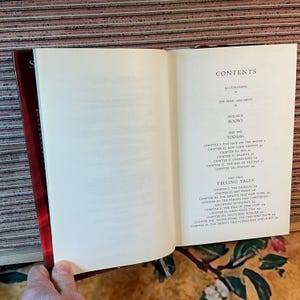 May include: Open book with a red cover, displaying the contents page. The text includes "CONTENTS," "ILLUSTRATIONS," and chapter titles like "The Face on the Water." The pages are cream-colored, and the book is held open by a hand.
