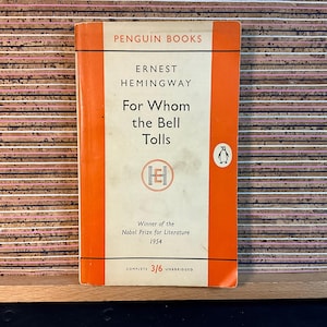 May include: A vintage Penguin Books paperback edition of "For Whom the Bell Tolls" by Ernest Hemingway. The cover is orange with white text and a penguin logo. The book was published in 1954 and is a complete and unabridged edition.