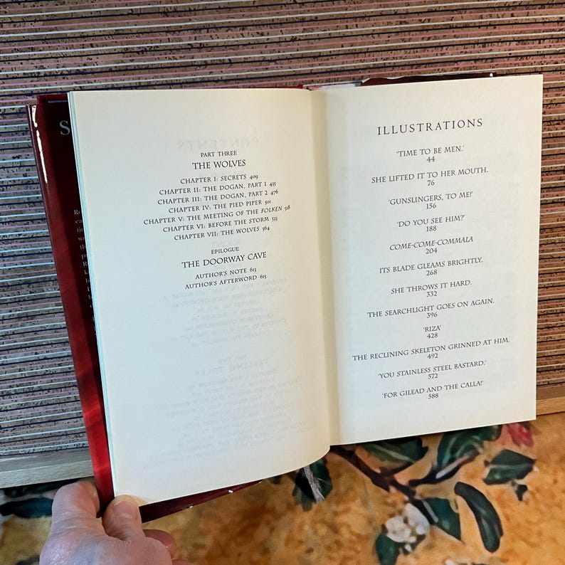 May include: Open book with red cover, displaying a table of contents and illustrations. The left page lists chapters like "The Wolves" and "The Doorway Cave." The right page shows illustrations with titles such as "Time to be Men" and "Gunslingers, to Me!"