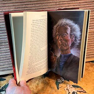 May include: An open book with a red cover, displaying an illustration of a man with white hair and a stern expression. The illustration is in color and appears to be a portrait. The book's pages are filled with printed text.