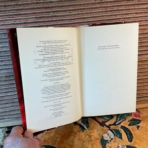 May include: An open book with white pages and red cover. The left page contains small text, while the right page has the text: "This book is for Frank Miller, who hears the voices in my head."