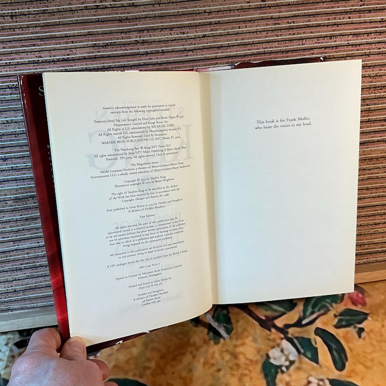 May include: An open book with white pages and red cover. The left page contains small text, while the right page has the text: "This book is for Frank Miller, who hears the voices in my head."