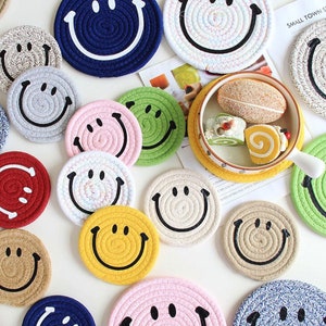 May include: A collection of colorful, circular coasters with smiley face designs. The coasters are made of woven rope and come in various colors, including blue, pink, green, and yellow. The coasters are approximately 4 inches in diameter.