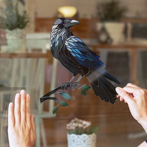 Puede incluir: Una calcomanía de cuervo estilo vitral con detalles azules y negros posada en una rama. La calcomanía se está aplicando a una ventana.