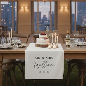 May include: A wedding reception table is set with a white table runner that reads "MR. & MRS. William 07.20.26". The table is decorated with a cake, candles, and wine glasses. Wooden chairs surround the table, and a chandelier hangs above.