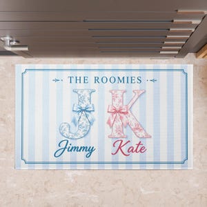 Könnte beinhalten: Eine rechteckige Fußmatte mit blau-weiß gestreiftem Hintergrund. Die Matte zeigt den Text "THE ROOMIES" und die Initialen "JK" in dekorativen floralen Mustern, mit den Namen "Jimmy" und "Kate" darunter.