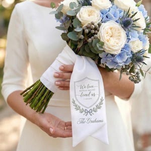 May include: A wedding bouquet with white and cream roses, blue hydrangeas, and foliage. The bouquet is tied with a white ribbon printed with the text "We love because He first loved us" and "The Grahams".