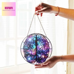 Puede incluir: Una ventana de vitral colgando de una cadena. La ventana es redonda y presenta una representación colorida de un cerebro humano con un diseño de galaxia. El cerebro es morado, azul y rosa con estrellas blancas.