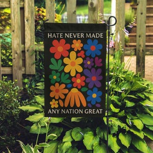May include: A black garden flag with the text "HATE NEVER MADE ANY NATION GREAT" above and below a colorful floral design. The flowers are in shades of red, orange, yellow, blue, and purple, with green leaves.