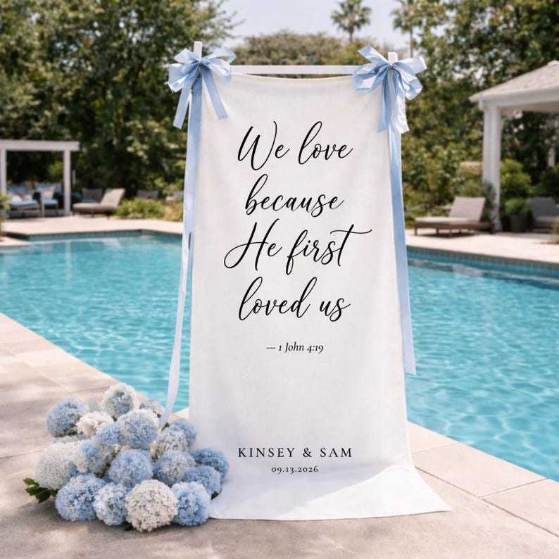 Puede incluir: Una pancarta de boda blanca con el texto "We love because He first loved us - 1 John 4:19" en escritura negra. La pancarta est&aacute; decorada con lazos azul claro y los nombres "KINSEY & SAM" y la fecha "09.13.2026".