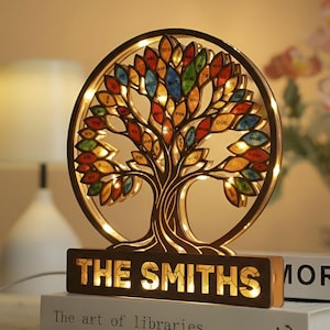 Puede incluir: Una lámpara de noche de árbol genealógico de madera con un marco circular y detalles de hojas coloridas. La base dice "THE SMITHS" en letras iluminadas. El diseño del árbol está intrincadamente cortado e iluminado con luces cálidas.