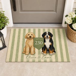 Puede incluir: Un felpudo rectangular con un patrón de rayas verticales en verde claro y crema. Presenta ilustraciones de un golden doodle y un boyero de Berna sosteniendo un banderín verde con el número 230, y el texto Olivia & Emma debajo.