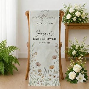 May include: A light-colored baby shower banner draped over a wooden chair. The banner features the text "A little wildflower is on the way" and "Jessica's Baby Shower 08.09.2026". Floral illustrations in muted tones adorn the banner.