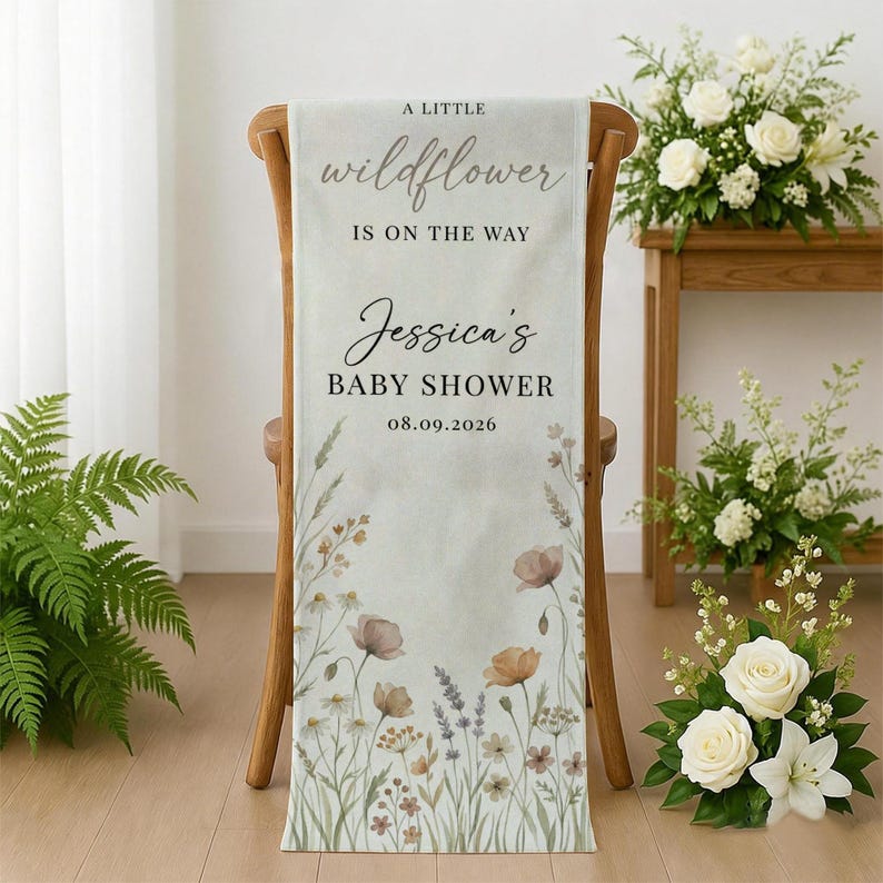 May include: A light-colored baby shower banner draped over a wooden chair. The banner features the text "A little wildflower is on the way" and "Jessica's Baby Shower 08.09.2026". Floral illustrations in muted tones adorn the banner.