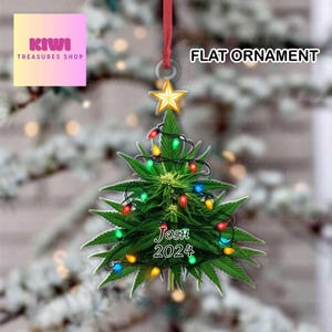 Op de afbeelding: Een platte kerstversiering in de vorm van een cannabisplant, versierd met kleurrijke lichtjes en een gouden ster. De versiering heeft de tekst "Josh 2024" en hangt aan een rood lint. De woorden "Flat Ornament" staan bovenaan.